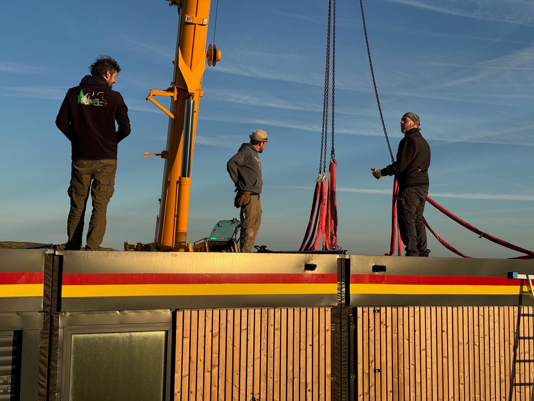 Trois compagnons debout sur la toiture d'un module pendant l'élingage, câbles de grue tendus, bandeau de rive coloré et bardage bois, ciel dégagé