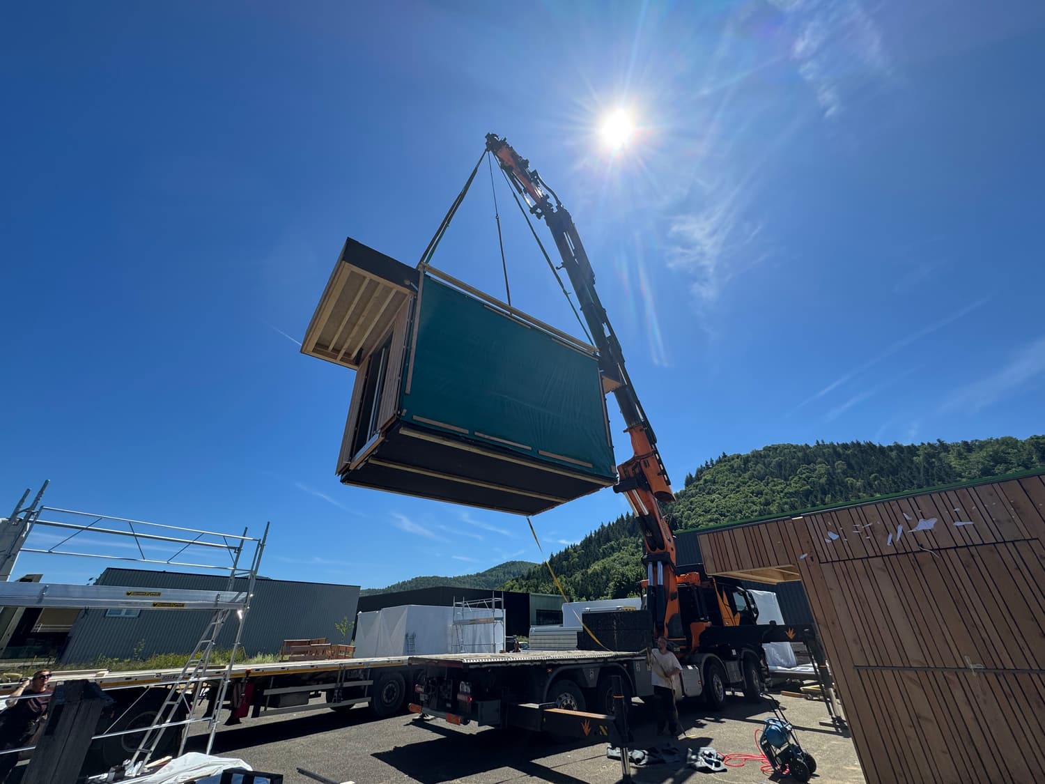 Module en ossature bois protégé par une bâche, levé par la grue d'un camion au-dessus d'une aire de chargement, collines boisées en arrière-plan