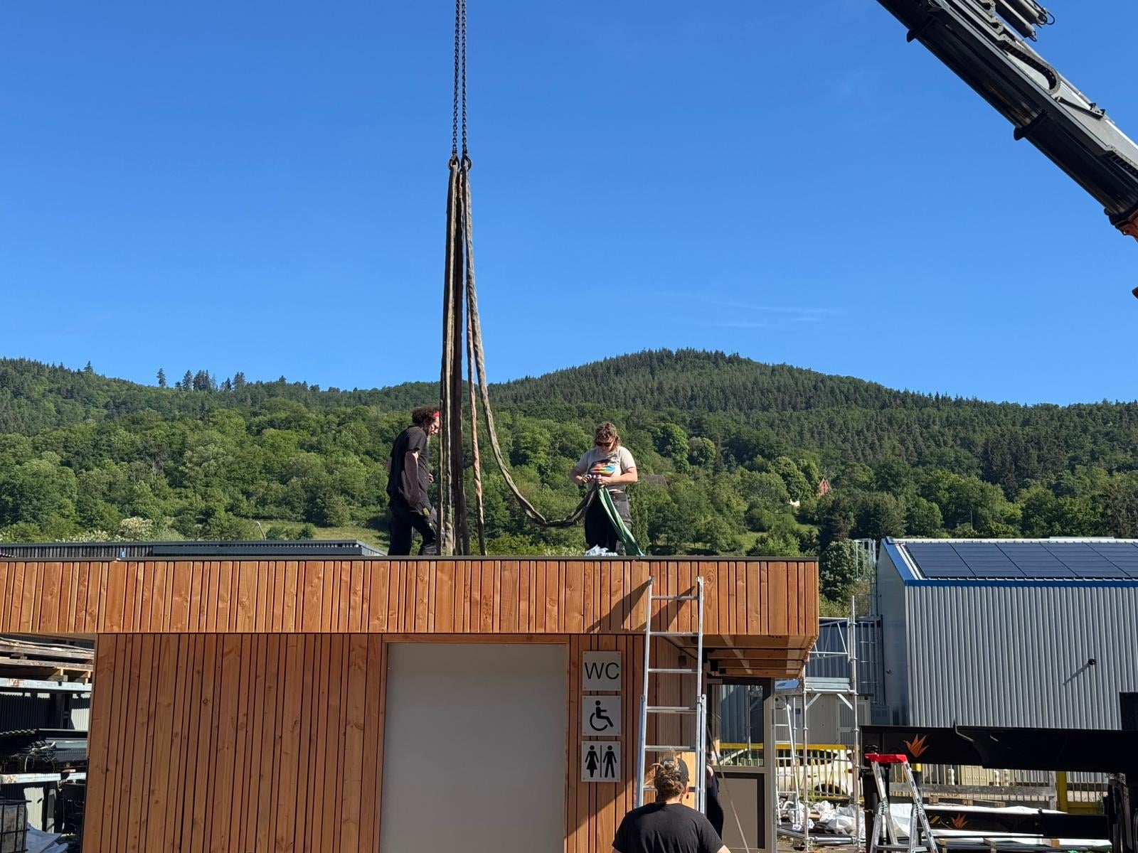 Deux compagnons sur la toiture d'un module fini, dans la cour de l'atelier, devant les collines de la vallée de Munster