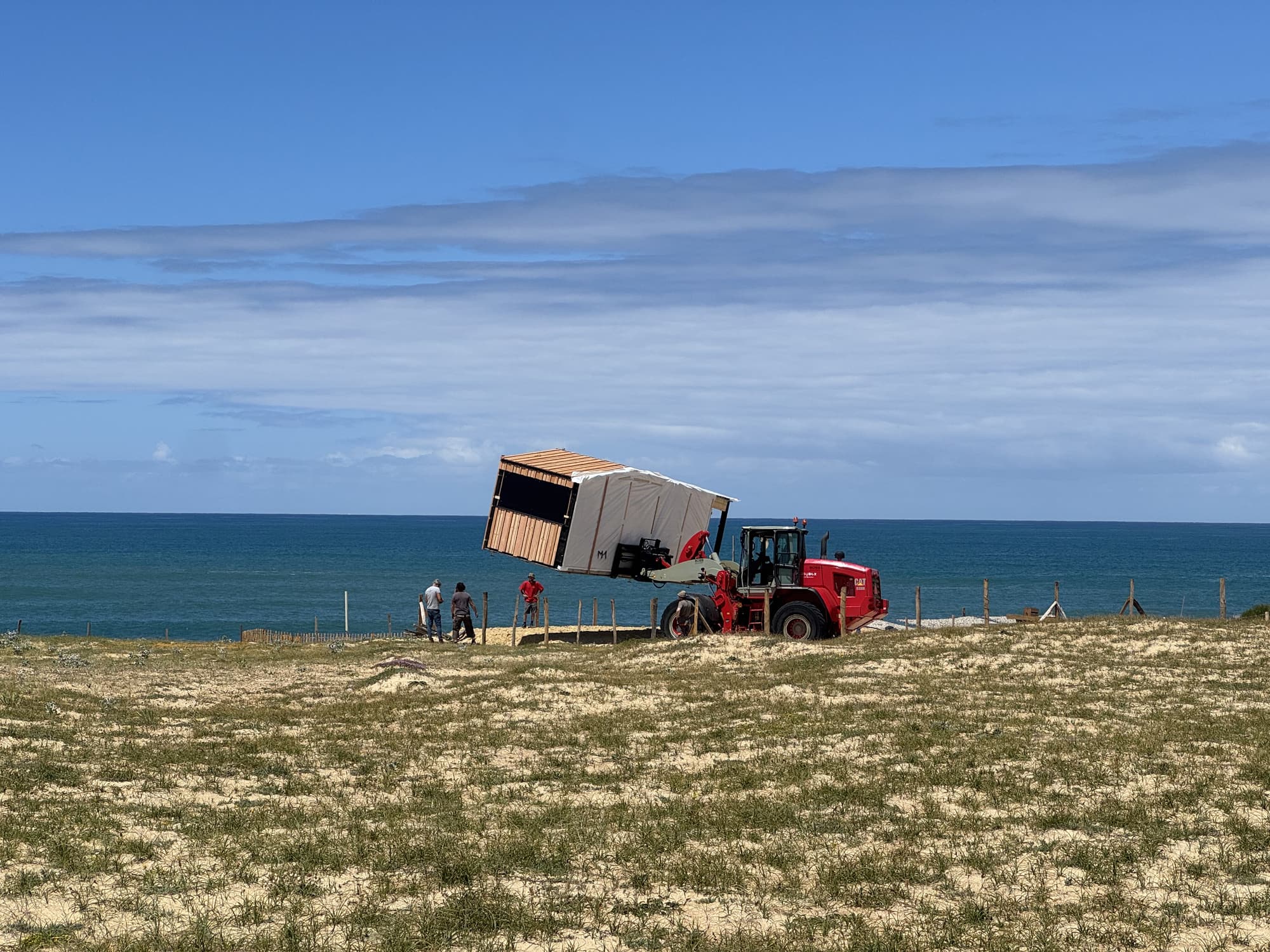 Module de poste de secours Hoummi soulevé par une chargeuse télescopique au sommet de la dune, face à l'océan, l'équipe de pose au sol