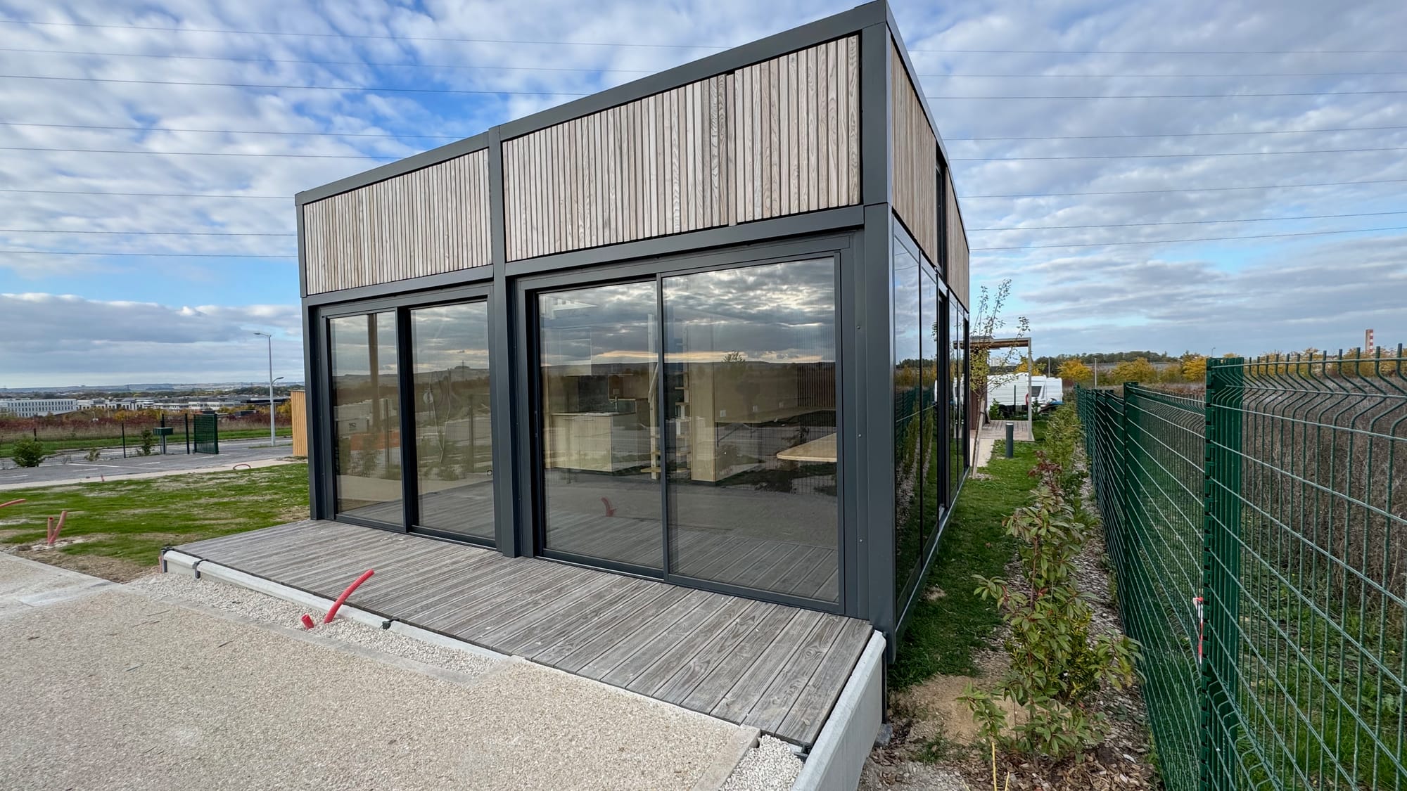 Module Hoummi posé sur son terrain, platelage de terrasse en cours de pose devant la façade vitrée, clôture et grue en second plan