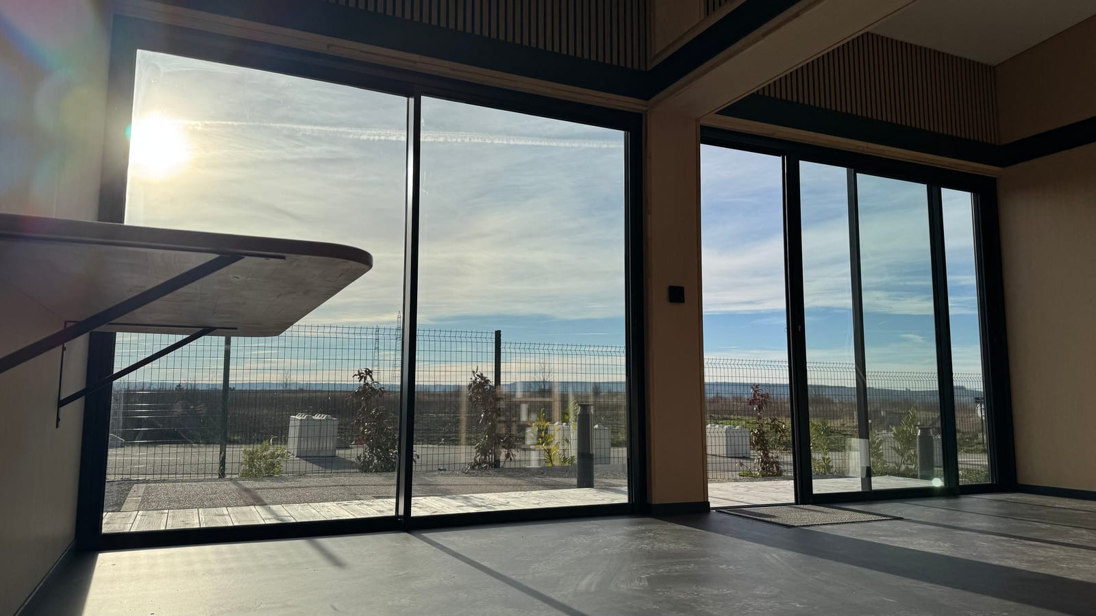 Plateau nu d'un module Hoummi à contre-jour : baie coulissante et angle vitré toute hauteur, sol béton, plafond à tasseaux